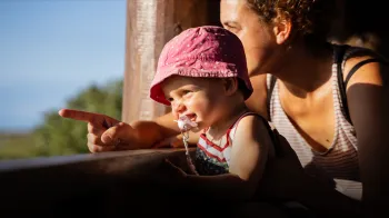 photo of a toddler smiling with pacifier looking into the distance with woman in the back pointing